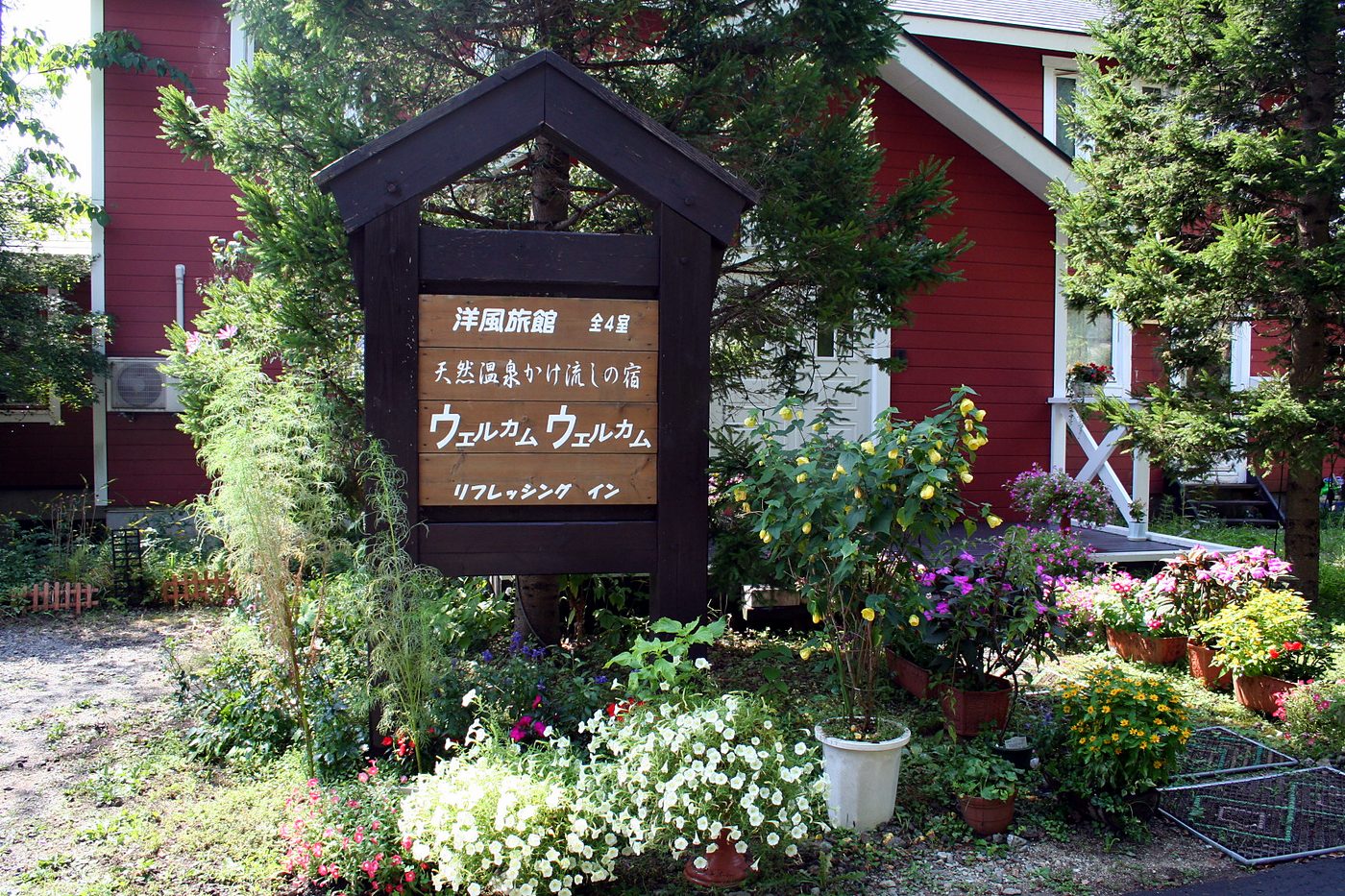 夏の玄関前。花に囲まれた看板と赤い壁の建物